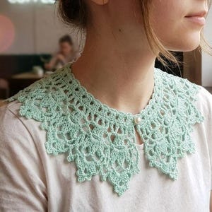 Puede incluir: Un cuello de ganchillo verde claro con un delicado dise&ntilde;o calado, abrochado con un solo bot&oacute;n de perla. El cuello se lleva sobre una camiseta blanca. El intrincado patr&oacute;n del cuello a&ntilde;ade un toque vintage al conjunto.