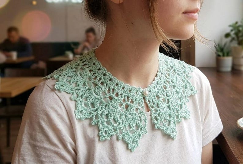Puede incluir: Un cuello de ganchillo verde claro con un delicado dise&ntilde;o calado, abrochado con un solo bot&oacute;n de perla. El cuello se lleva sobre una camiseta blanca. El intrincado patr&oacute;n del cuello a&ntilde;ade un toque vintage al conjunto.