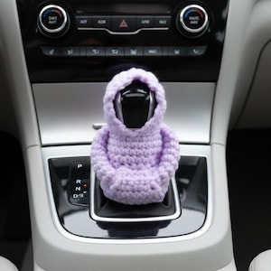 Könnte beinhalten: Ein lavendelfarbener gehäkelter Schalthebelbezug in Form eines Kapuzenpullovers. Der Bezug wird über den Schalthebel eines Autos gelegt, wobei die Kapuze hochgeklappt ist. Der Innenraum des Autos hat eine silberne Konsole und schwarze Bedienelemente.