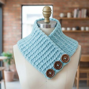May include: A light blue crocheted cowl with a ribbed texture, displayed on a mannequin. The cowl features three large, round, brown wooden buttons on the front. The background includes a brick wall and wooden shelves.