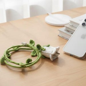 Op de afbeelding: Een groene gehaakte kabel met bladdetails en een witte gehaakte stekker. Een witte smartphone met een doorzichtige hoes ligt op een stapel boeken. De items liggen op een houten oppervlak.