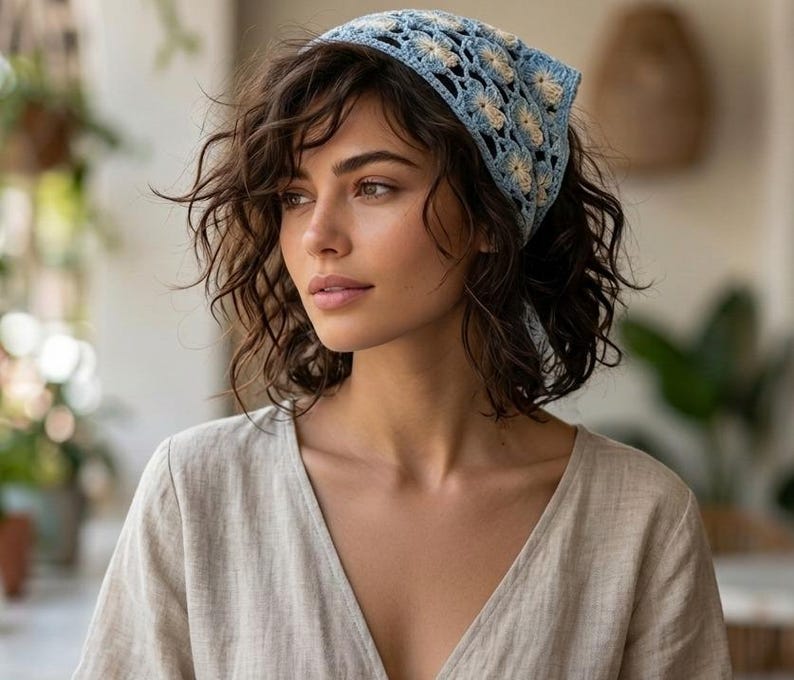 May include: A light blue and cream-colored crochet headscarf with a floral pattern. The headscarf is tied at the back. The person is wearing a beige v-neck top.