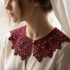 Puede incluir: Un cuello de ganchillo burdeos con borde festoneado y cierre de botón de perla. El cuello se lleva sobre una blusa blanca. El intrincado diseño de encaje añade un toque vintage a la prenda.
