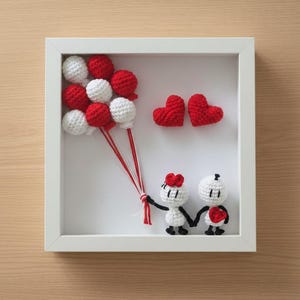 May include: A framed crochet artwork showing a couple holding hands, with a heart and balloons. The balloons are red and white, and the couple is black and white. The frame is white.