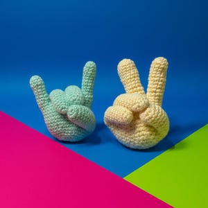 Könnte beinhalten: Zwei gehäkelte Handgesten: eine in Hellblau, die das 'Ich liebe dich'-Zeichen bildet, und die andere in Gelb, die das Friedenszeichen zeigt. Vor einem lebendigen Hintergrund aus Blau, Pink und Grün.
