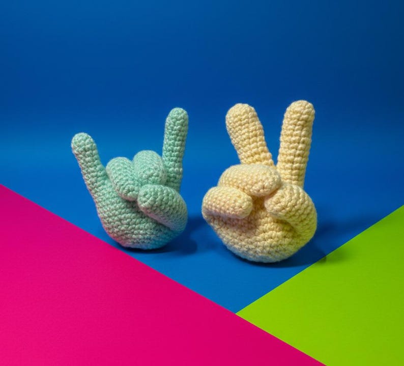 May include: Two crocheted hand gestures: one in light blue, forming the 'I love you' sign, and the other in yellow, displaying the peace sign. Set against a vibrant backdrop of blue, pink, and green.