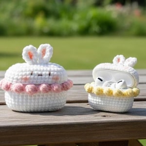 May include: Two white crochet cases, one closed and one open, shaped like cupcakes with bunny ears. The closed case has a pink trim, while the open case has a yellow trim. The open case reveals white earbuds inside.