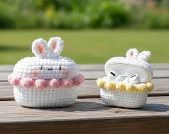 Pode incluir: Duas capas brancas de crochê, em forma de cupcakes com orelhas de coelho. Uma está fechada com uma borda rosa, a outra aberta com uma borda amarela. A capa aberta revela fones de ouvido brancos.