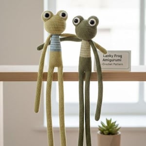 May include: Two handmade crochet frog amigurumi toys. One frog is light green with a blue and white striped shirt, the other is dark green with a white and tan striped shirt. The frogs have long legs and large eyes. A sign reads "Lanky Frog Amigurumi - Crochet Pattern."