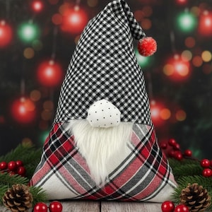 Pode incluir: Um gnomo de Natal decorativo com um chapéu xadrez preto e branco e um pompom vermelho. O gnomo tem barba branca e corpo xadrez vermelho, preto e branco. Está sobre uma superfície de madeira com pinhas e bagas vermelhas.