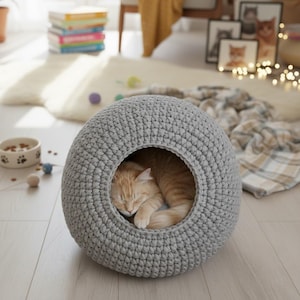 May include: A grey, crocheted cat bed in a round shape. An orange tabby cat is asleep inside the bed. The bed is on a light wood floor. Cat toys and a food bowl are nearby.