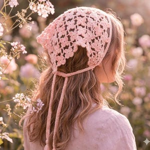 May include: A light pink, crocheted bonnet with a floral pattern. The bonnet has long ties and a tassel at the end of each tie. The bonnet is worn over long, wavy hair. The background is a soft focus of flowers.
