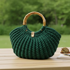 Può includere: Borsa a mano all'uncinetto verde scuro fatta a mano con manico in bambù. La borsa presenta un design a coste e una piccola etichetta in pelle. La borsa è su una superficie di legno.