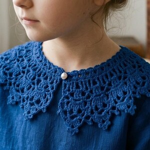 Puede incluir: Un cuello de crochet azul con borde festoneado y cierre de bot&oacute;n de perla. El cuello se lleva sobre una camisa azul. El crochet tiene un dise&ntilde;o abierto y calado. La imagen es un primer plano del cuello y la camisa.