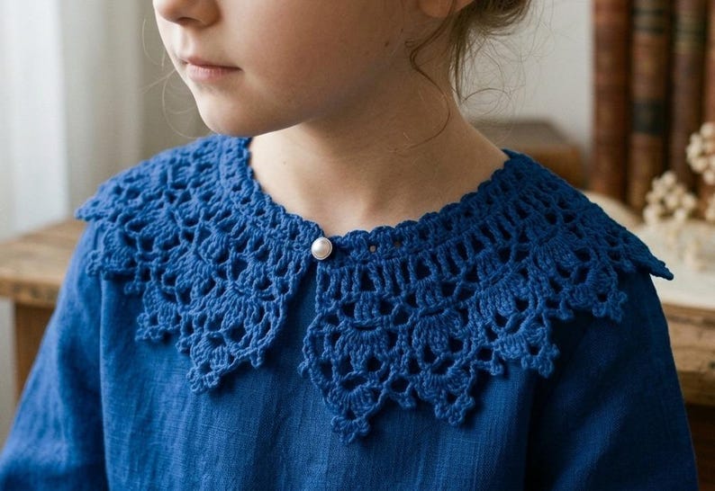 Puede incluir: Un cuello de crochet azul con borde festoneado y cierre de bot&oacute;n de perla. El cuello se lleva sobre una camisa azul. El crochet tiene un dise&ntilde;o abierto y calado. La imagen es un primer plano del cuello y la camisa.