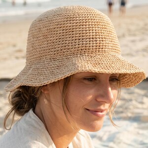 Puede incluir: Un sombrero de sol beige tejido a ganchillo con un ala ligeramente acampanada. El sombrero está hecho de un material tejido y tiene una copa redondeada. El sombrero es adecuado para la playa o actividades al aire libre.