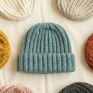 May include: A collection of six ribbed knit beanies in various colours including cream, beige, teal, mustard yellow, burnt orange, dusty pink, and charcoal grey. Each beanie features a thick folded cuff and a vertical knit pattern throughout the fabric.