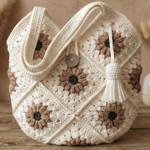 May include: Cream-colored crochet tote bag with a floral pattern in brown and dark green. The bag features a button closure, a tassel, and a shoulder strap. The bag is sitting on a wooden surface.