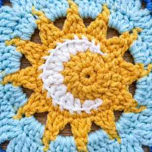 Puede incluir: Un motivo de ganchillo cuadrado que presenta un sol amarillo central con una luna creciente blanca superpuesta. El sol está rodeado por puntadas de hilo azul claro, todo enmarcado por un borde azul oscuro. El diseño es de hilo texturizado.