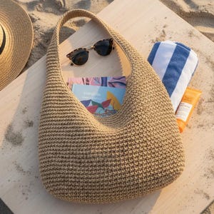 Puede incluir: Bolso de hombro beige tejido a ganchillo con forma curva, que contiene un libro y gafas de sol. Una toalla a rayas azules y blancas y un tubo de protector solar están junto al bolso. Un sombrero de paja es parcialmente visible en el fondo. Los artículos están sobre una superficie de madera.