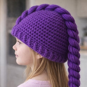 Può includere: Un berretto all'uncinetto color viola vivace caratterizzato da una spessa coda di lana intrecciata che si estende dalla corona fino alla schiena. Il cappello presenta un motivo a maglia testurizzato e un bordo a coste in filato viola.