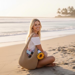 May include: A beige hobo-style shoulder bag with a textured, woven knit construction. The bag features a single wide strap and a spacious open-top design, containing a small patterned book and a bright yellow sunflower tucked into the side.