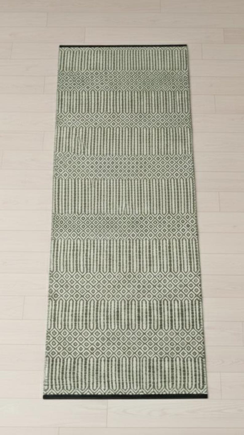 Puede incluir: Una alfombra de pasillo rectangular con un patr&oacute;n geom&eacute;trico repetitivo en verde salvia y blanco roto. El dise&ntilde;o consiste en bandas horizontales alternas de diamantes y formas ovaladas alargadas, con un borde negro s&oacute;lido en ambos extremos.