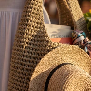 May include: A beige crocheted tote bag with a thick shoulder strap, containing a book and a patterned silk scarf, paired with a matching beige straw hat featuring a thin black ribbon band around the crown, all captured in warm sunlight.