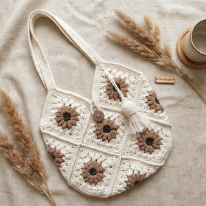 May include: A crochet tote bag constructed from cream and brown floral granny squares. The bag features a single shoulder strap, a central wooden button closure, and a cream-coloured tassel accent. A small wooden tag labelled Handmade is attached.