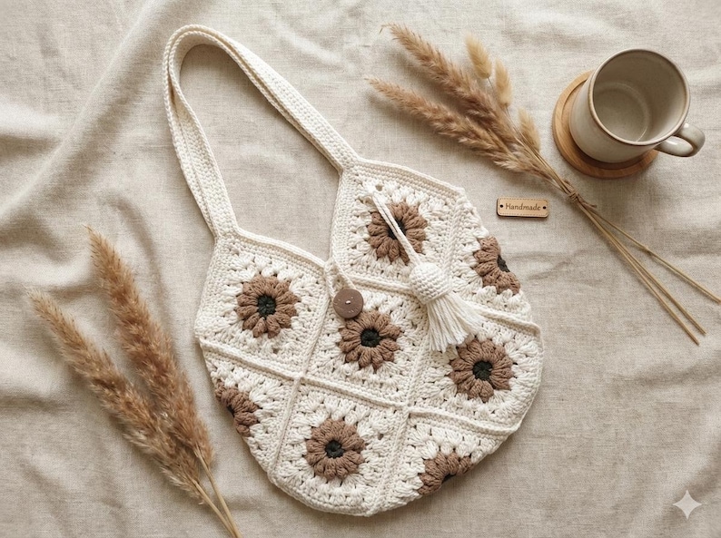 May include: A crochet tote bag constructed from cream and brown floral granny squares. The bag features a single shoulder strap, a central wooden button closure, and a cream-coloured tassel accent. A small wooden tag labelled Handmade is attached.