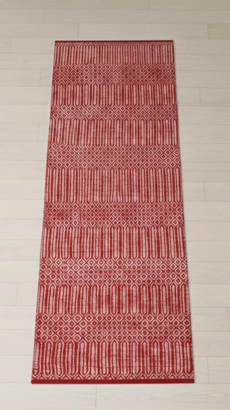 Puede incluir: Una alfombra de pasillo rectangular con un vibrante patr&oacute;n geom&eacute;trico en rojo y blanco. El dise&ntilde;o consiste en bandas horizontales alternas de formas de diamante intrincadas y motivos verticales alargados, creando una textura sim&eacute;trica.