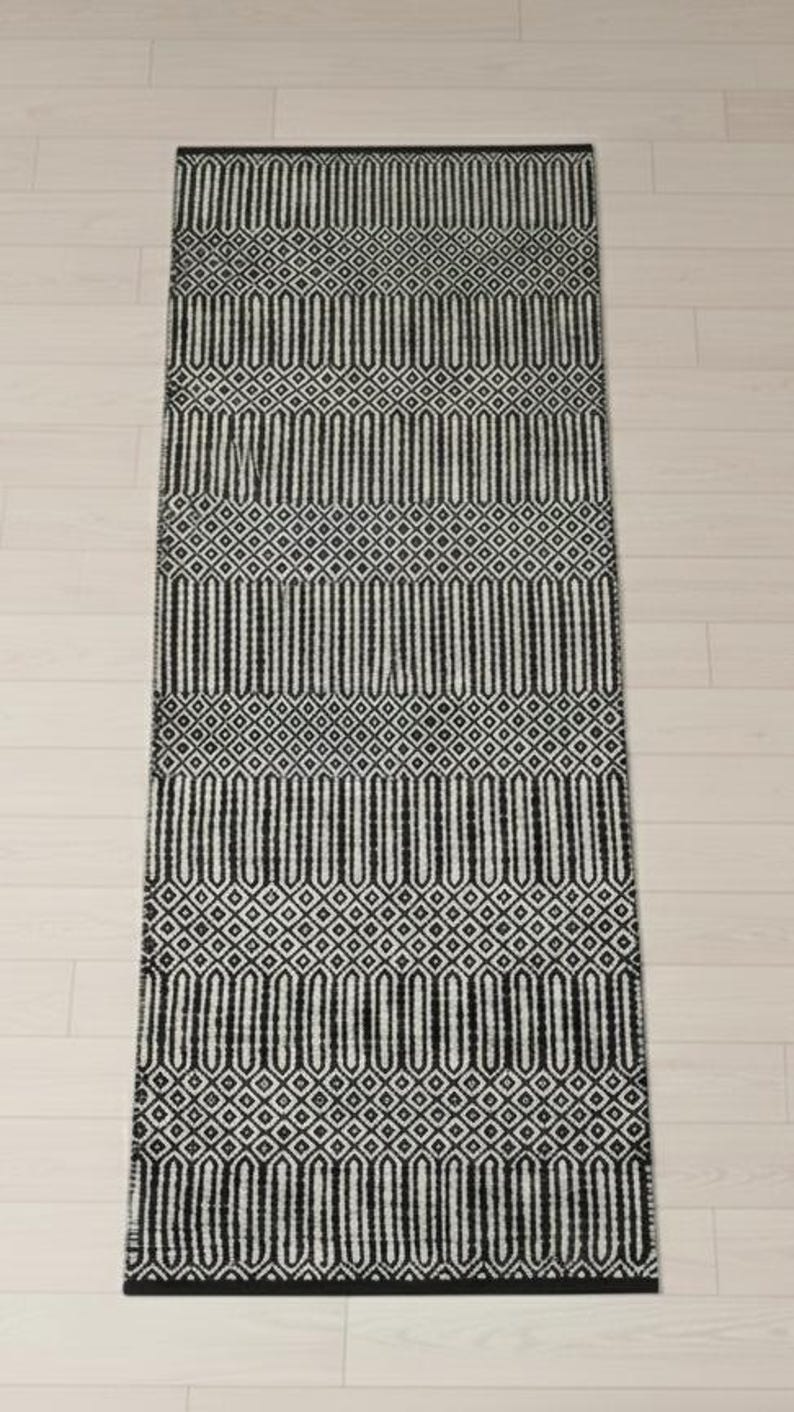 Puede incluir: Una alfombra de pasillo rectangular con un patr&oacute;n geom&eacute;trico repetitivo en blanco y negro. El dise&ntilde;o consta de bandas horizontales alternas de formas ovaladas verticales alargadas y peque&ntilde;as cuadr&iacute;culas de rombos, con un borde negro s&oacute;lido.