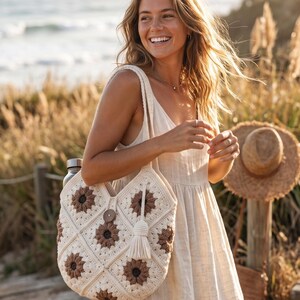 May include: A cream-coloured crochet tote bag featuring a repeating pattern of brown and cream sunflower-style motifs. The bag includes a single wooden button closure at the top centre and a long, cream-coloured yarn tassel hanging from the side strap.