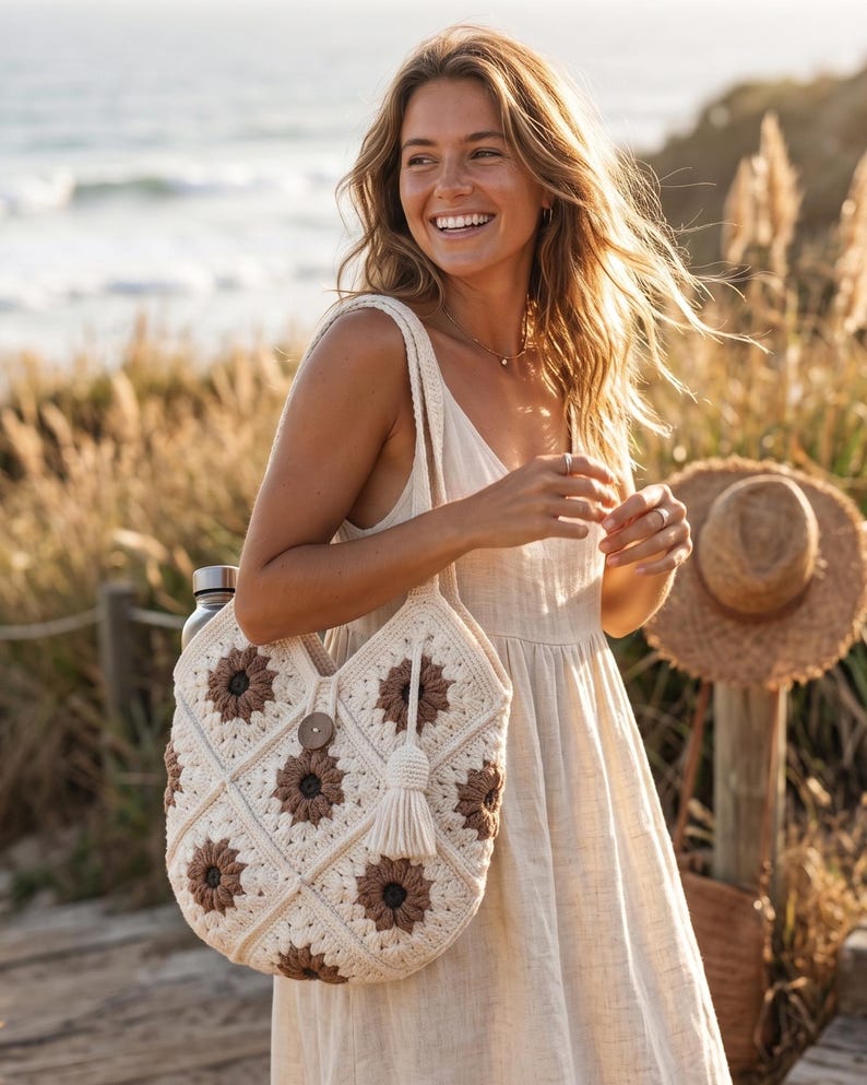 May include: A cream-coloured crochet tote bag featuring a repeating pattern of brown and cream sunflower-style motifs. The bag includes a single wooden button closure at the top centre and a long, cream-coloured yarn tassel hanging from the side strap.