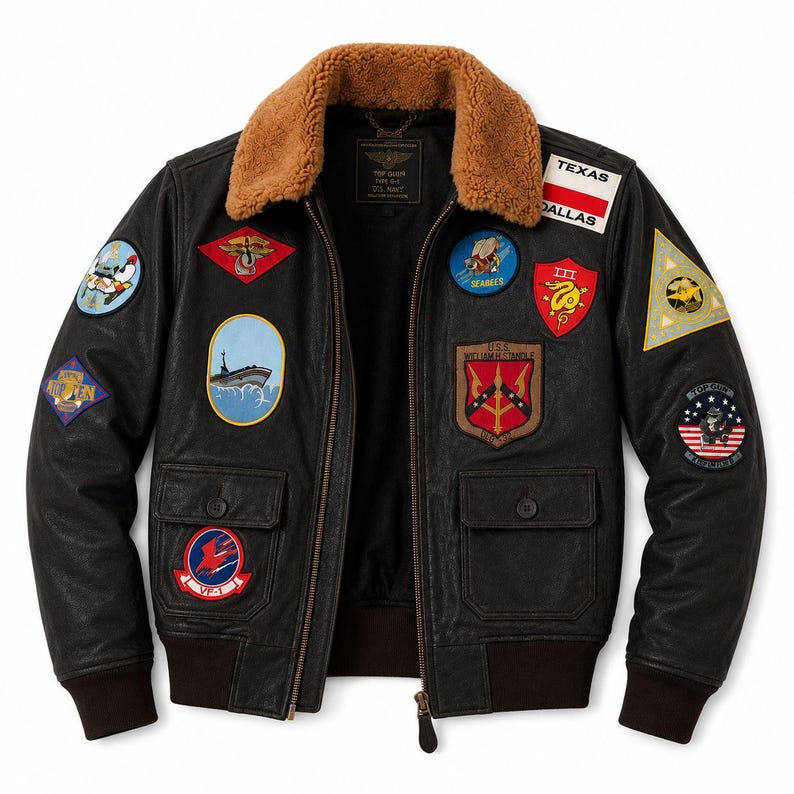 Peut inclure: Un blouson aviateur en cuir noir avec un col en fausse fourrure marron, une fermeture &eacute;clair frontale et deux poches &agrave; rabat. La veste est orn&eacute;e de nombreux &eacute;cussons brod&eacute;s de style militaire, incluant les textes Texas, Dallas et U.S.S. William H. Standley.