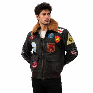 Peut inclure: Un blouson aviateur en cuir noir avec un col en fausse fourrure marron, une fermeture &eacute;clair frontale et deux poches &agrave; rabat. La veste est orn&eacute;e de divers &eacute;cussons brod&eacute;s color&eacute;s sur la poitrine et les manches, incluant du texte et des motifs.