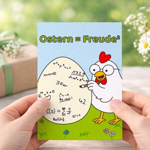 Könnte beinhalten: Eine von Händen gehaltene Grußkarte zeigt ein Cartoon-Huhn mit Brille, das einen Bleistift in der Nähe eines großen Eis mit mathematischen Gleichungen hält. Die Karte lautet "Ostern = Freude²" in grünen Buchstaben. Eine Geschenkbox und Gänseblümchen sind im Hintergrund.