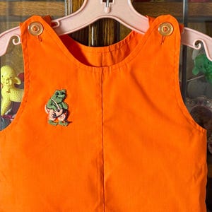 May include: Bright orange children's overalls with a frog applique. The overalls have a classic bib design with button closures on the straps. The frog applique is green and wearing pink pants. The overalls are hanging on a pink hanger.