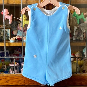 May include: A light blue baby onesie with white trim and a white anchor and snowflake design. The onesie is hanging on a wooden hanger.