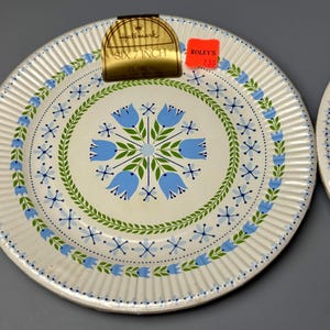 May include: Two white, 7-inch plates with a floral design. The plates feature a cream-colored center with blue tulip-like flowers and green leaves. The rim has a ribbed texture and is decorated with blue and green accents.