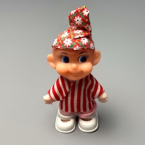 May include: A vintage doll with a smiling face, blue eyes, and a red and white striped shirt. The doll wears a red hat with a white floral pattern and white shoes. The doll is standing on a gray surface.