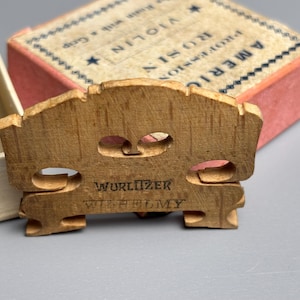 May include: A wooden violin bridge with the text "Wurlitzer Wilhelmy" is in the foreground. A box of American Professional Rosin is in the background. The bridge is light brown with two circular cutouts.