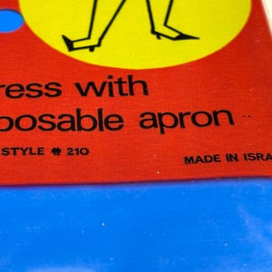 May include: Close-up of a product label with the words "dress with disposable apron" in black text on a red background. The label also includes "STYLE # 210" and "MADE IN ISRAEL". A blue circle and a yellow circle with a black illustration are visible.
