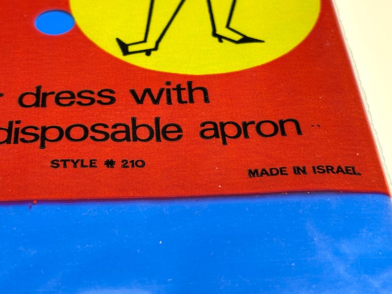 May include: Close-up of a product label with the words "dress with disposable apron" in black text on a red background. The label also includes "STYLE # 210" and "MADE IN ISRAEL". A blue circle and a yellow circle with a black illustration are visible.