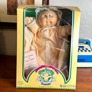 May include: A Cabbage Patch Kids doll in a pink and white gown with a green and yellow box. The box says "1985 Cabbage Patch Kids Preemie! I'm unique, I'm adoptable." and has the March of Dimes logo.