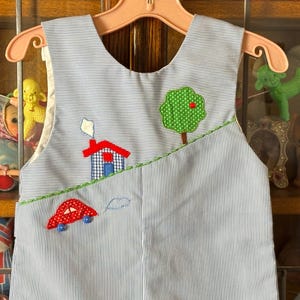 May include: A blue and white striped toddler's bib overall with appliqued designs. The bib features a red car, a house, a tree, and a cloud. The overall is hanging on a pink hanger.
