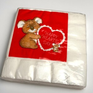 May include: A stack of white paper napkins wrapped in clear plastic. The top napkin features a red square with a cartoon koala holding a heart that says "HAPPY HEARTS' DAY!" A small mouse is also on the heart.