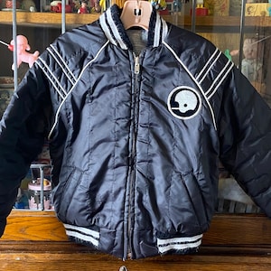 May include: A black satin bomber jacket with white piping and a white football helmet graphic on the left chest. The jacket has a zipper closure and a ribbed collar, cuffs, and hem.