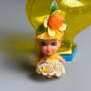 May include: A vintage doll with yellow hair, blue eyes, and a floral headpiece. The doll wears a yellow dress with a lace overlay. A yellow, flower-shaped container is in the background.