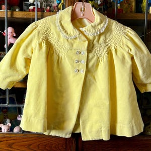 May include: A yellow corduroy jacket with a white lace collar and button closure. The jacket has a smocked bodice and is perfect for a little girl.